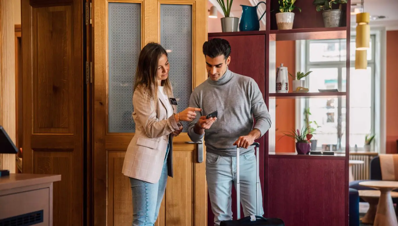 Host zeigt einem Gast mit Koffer etwas auf dem Smartphone und unterstützt ihn beim Ankommen im Hotel.