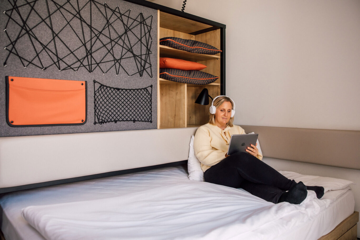 Frau sitzt entspannt auf einem Bett mit Tablet und Kopfhörern vor Wandmodul mit Netzstruktur und Stauraum.