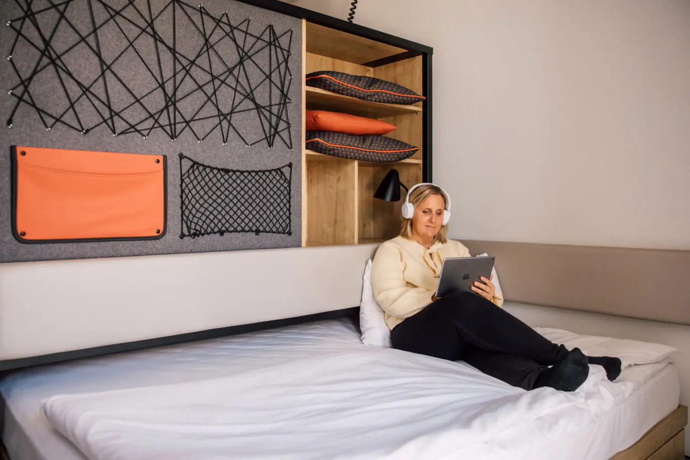 A woman sits relaxed on a bed with a tablet and headphones in front of a wall module with net storage.