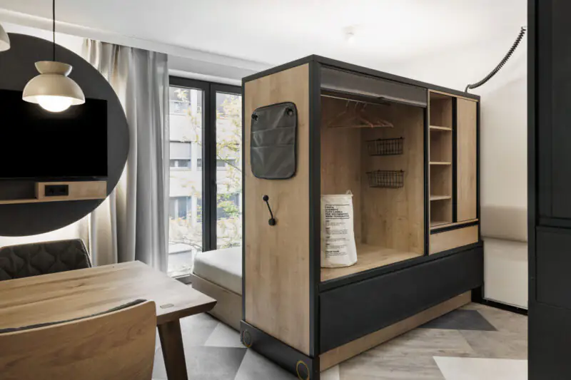 Room divider as open wardrobe with shelves and wood details separates living and sleeping area.