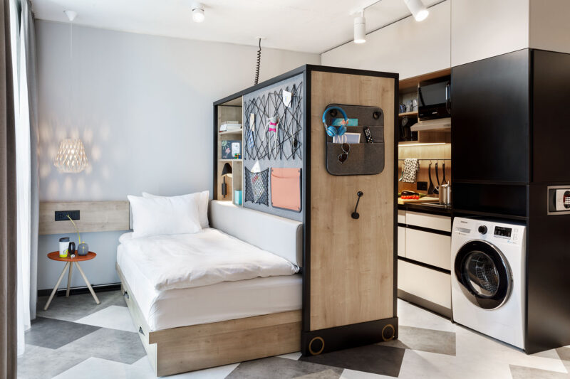 Helles Studio mit Bett, Netzelement, Sessel und Küche im Hintergrund – modernes Raumdesign mit warmen Farben.