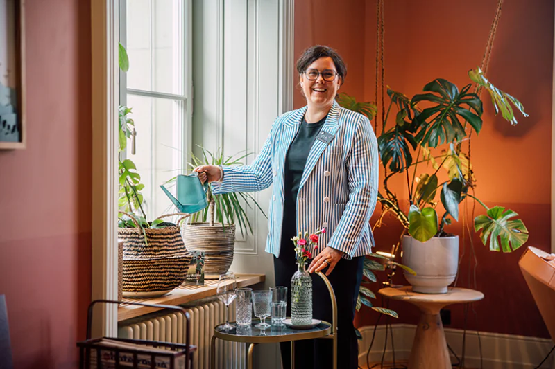 Smiling hotel host watering plants in a warm, plant-filled lounge.