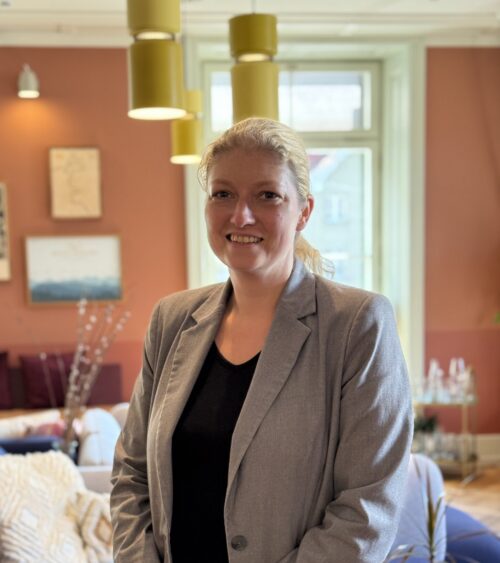 Host Andrea steht lächelnd in einem gemütlichen Stay KooooK Gemeinschaftsbereich mit Sofa und farbenfrohem Interior.