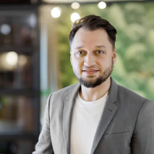Portrait von Host Alex mit Lächeln im grauen Sakko vor grüner Wand mit dekorativen Lichtelementen.