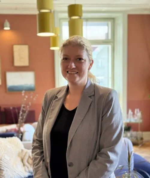 Host Andrea steht lächelnd in einem gemütlichen Stay KooooK Gemeinschaftsbereich mit Sofa und farbenfrohem Interior.