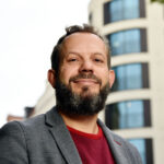 Mann im Business-Outfit vor Gebäude mit Rundturm, seitlich von vorne fotografiert.